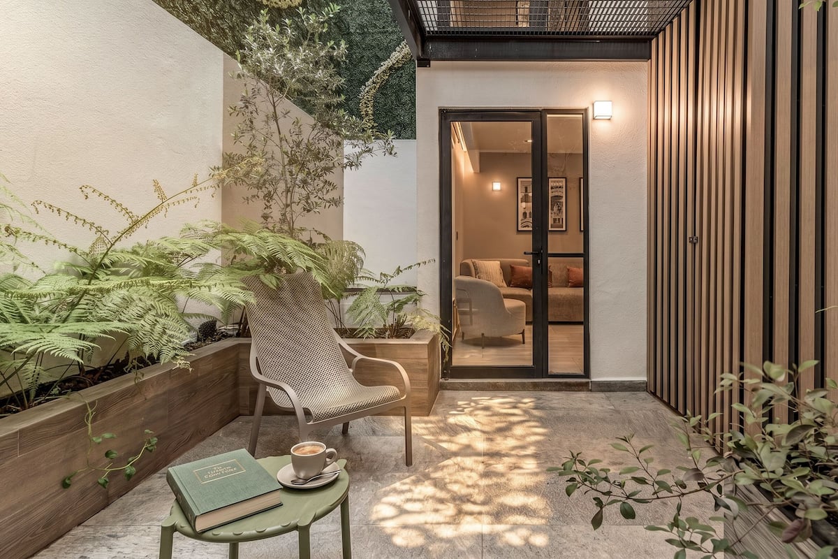 Un rincón exterior acogedor con una silla, una mesa con un libro y una taza, y una maceta de madera con helechos.