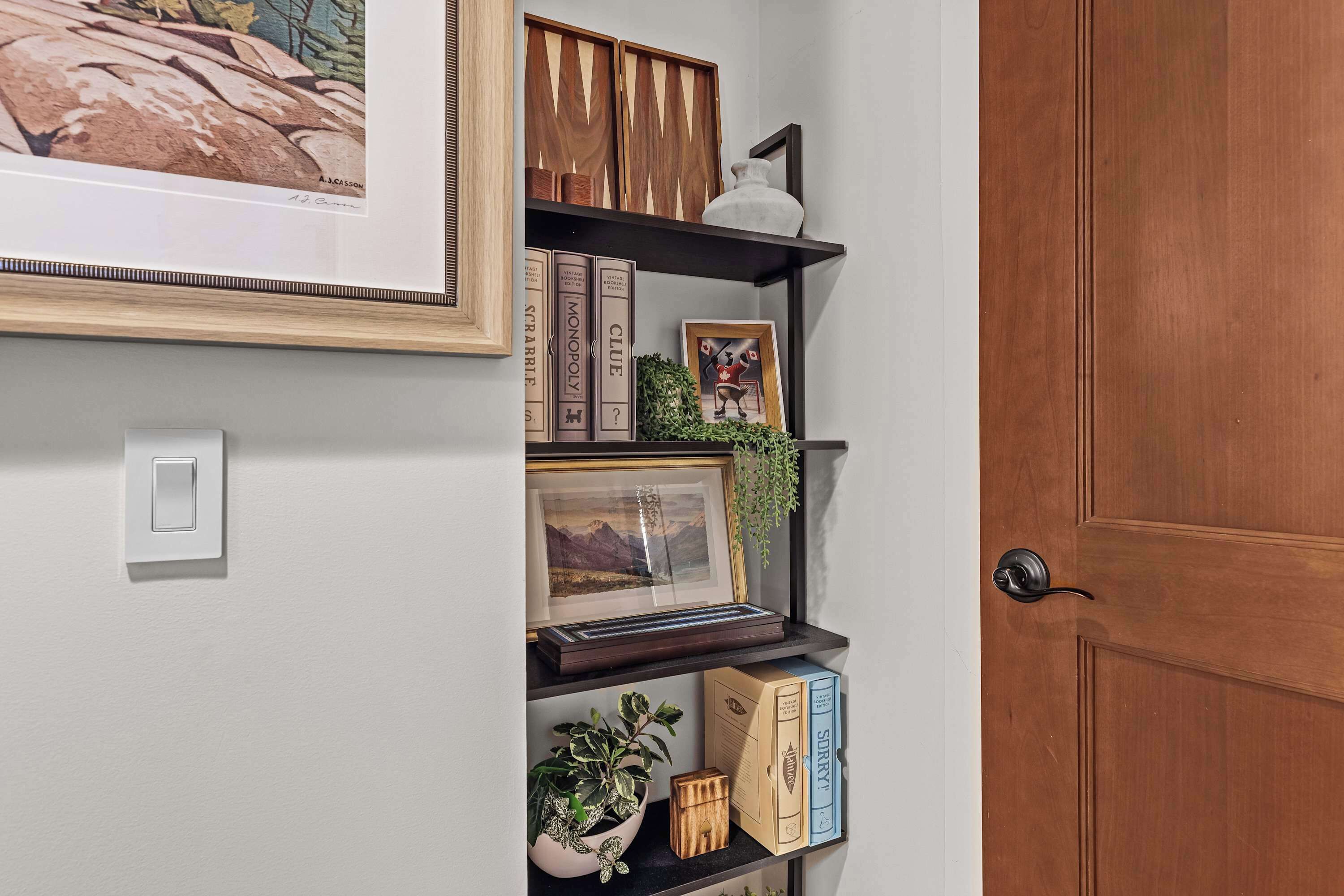 A hallway with a wooden door, a framed picture, a shelf with books and decorative items, and a potted plant.
