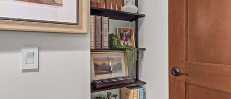 A hallway with a wooden door, a framed picture, a shelf with books and decorative items, and a potted plant.