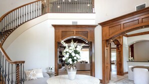 A grand foyer with a marble floor, a chandelier, a round table with a vase of flowers, and a staircase with wooden railings.