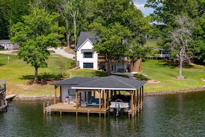 A lakeside property with a dock, a covered seating area, and a two-story house with a deck.