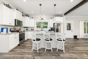 A kitchen with white cabinets, a stainless steel refrigerator, a microwave, and a window with greenery outside. There are three white bar stools and a countertop with a sink.