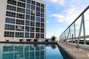 A hotel pool with a view of a high-rise building, lounge chairs, and a glass railing.