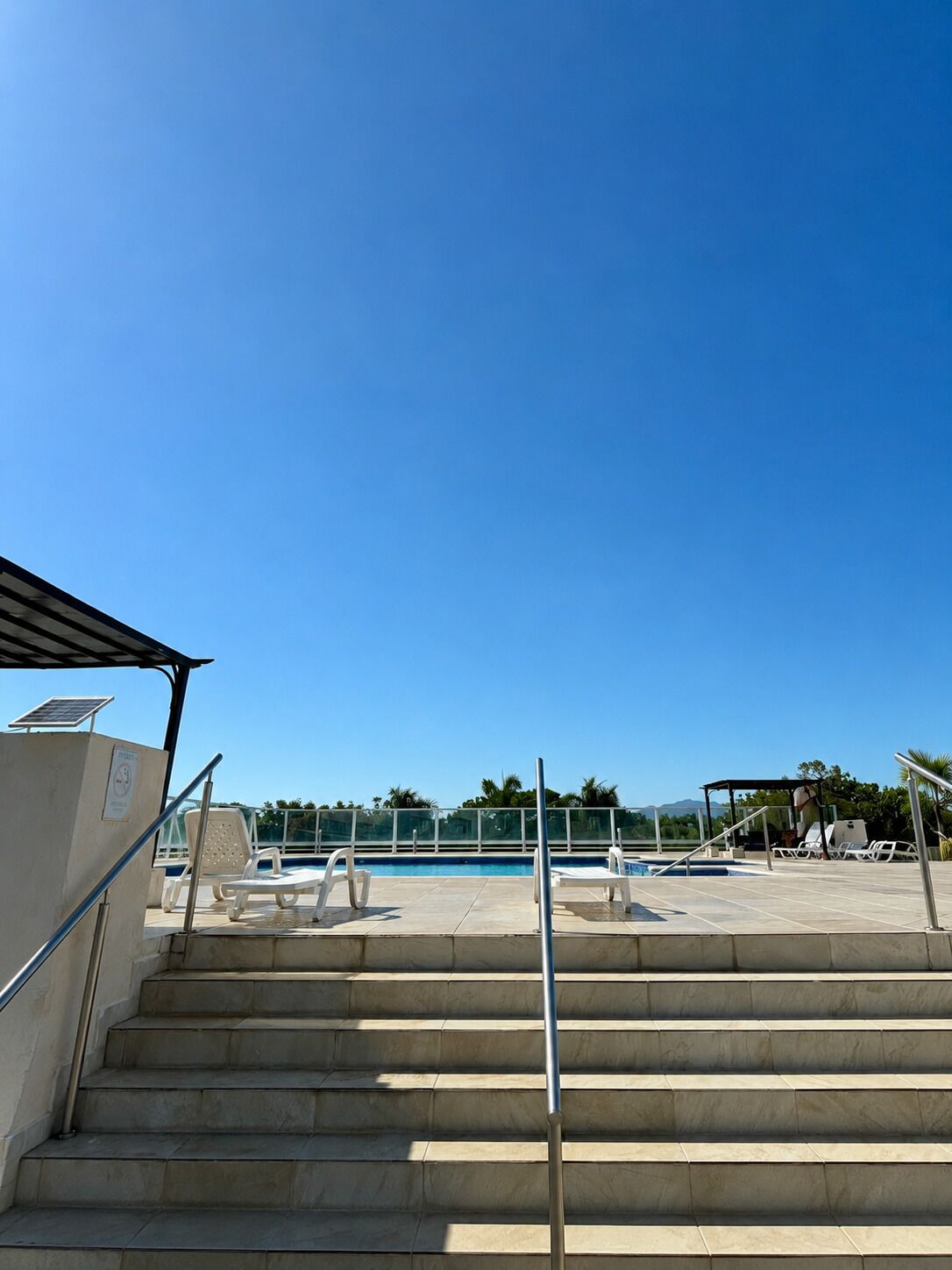 Ein Poolbereich mit Stufen zum Wasser, einem überdachten Sitzbereich und wolkenlosem, blauem Himmel.