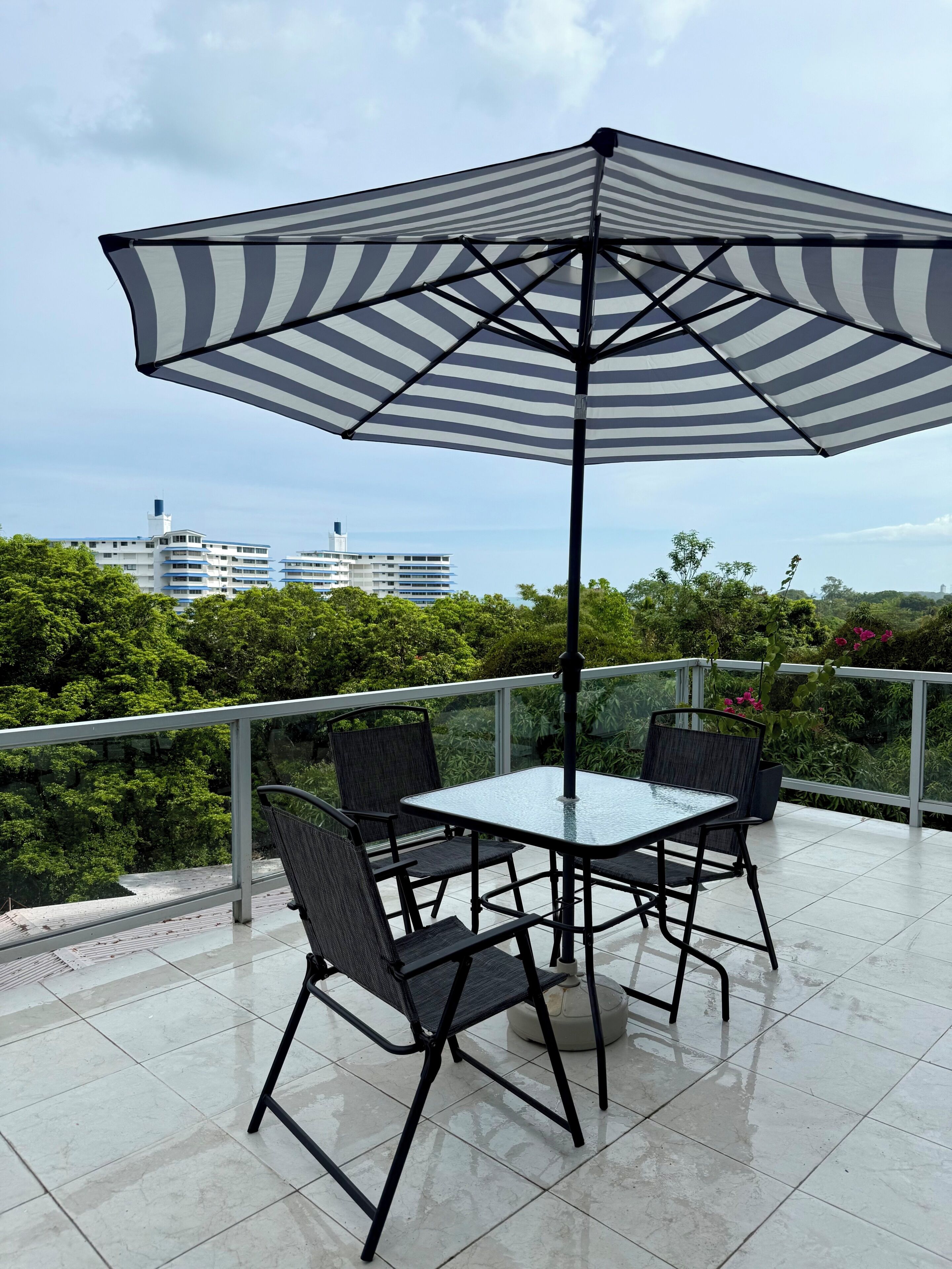 Un patio con una mesa y sillas, un parasol a rayas, y vistas a edificios y vegetación.