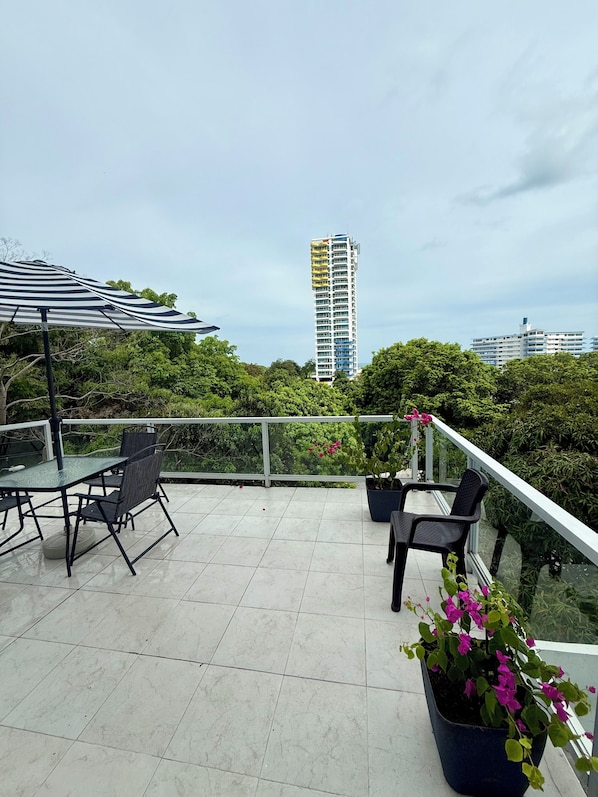 Una terraza en la azotea con barandilla de vidrio, mobiliario exterior y vista a un edificio de gran altura.