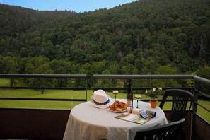 Ein Tisch mit Hut, Brezeln, Weingläsern und einem Buch auf einem Balkon mit Blick auf einen bewaldeten Bereich.