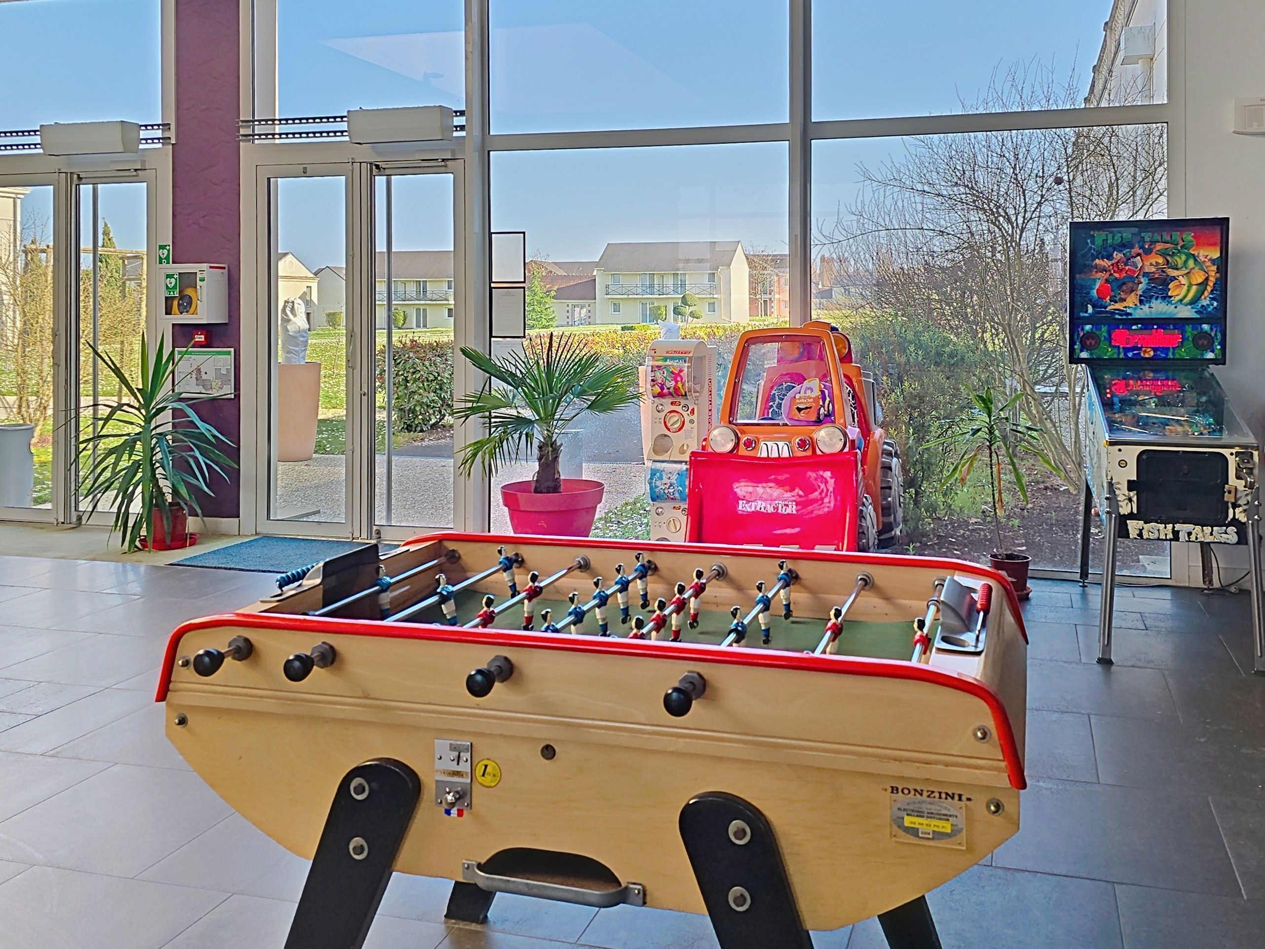 Ein Tischkicker in einem Raum mit großen Fenstern, einem Feuerlöscher und einem Fernseher, der ein Spiel zeigt.