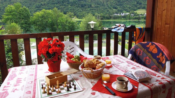 Een tafel gedekt met eten, drinken en een schaakbord op een balkon met uitzicht op een meer en heuvels.
