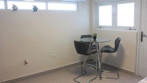 Una habitación pequeña con una mesa redonda de vidrio, dos sillas negras, una planta en maceta, una ventana con persianas, un equipo de aire acondicionado y una puerta.