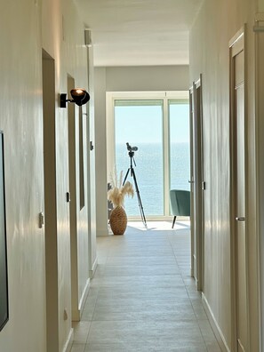 Ein Flur mit einer Kamera auf einem Stativ, einer Vase mit getrockneten Pflanzen und Blick auf das Meer durch große Fenster.