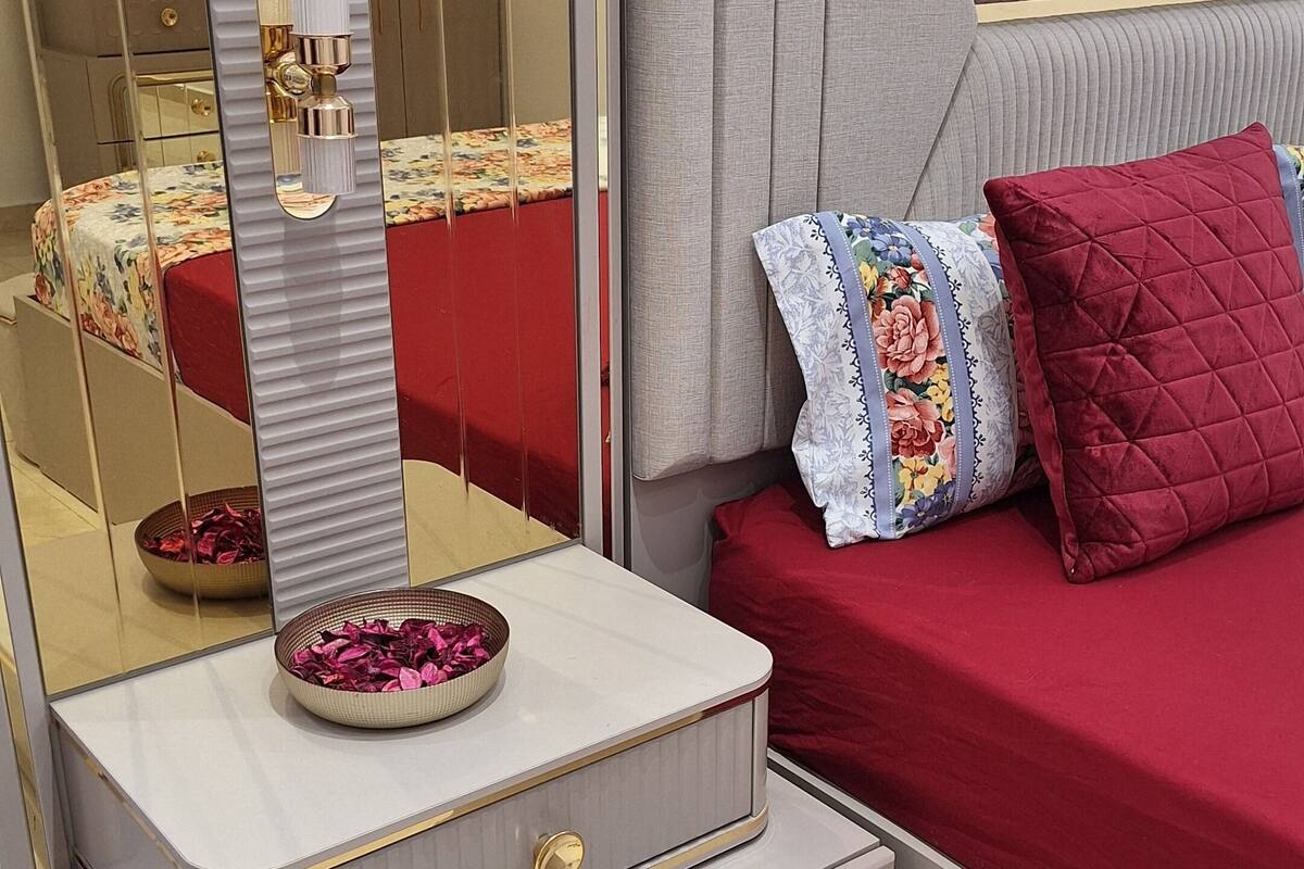Un dormitorio con un espejo grande, una tocador con un jarrón de flores, una cama con ropa de cama roja y un cabecero gris.
