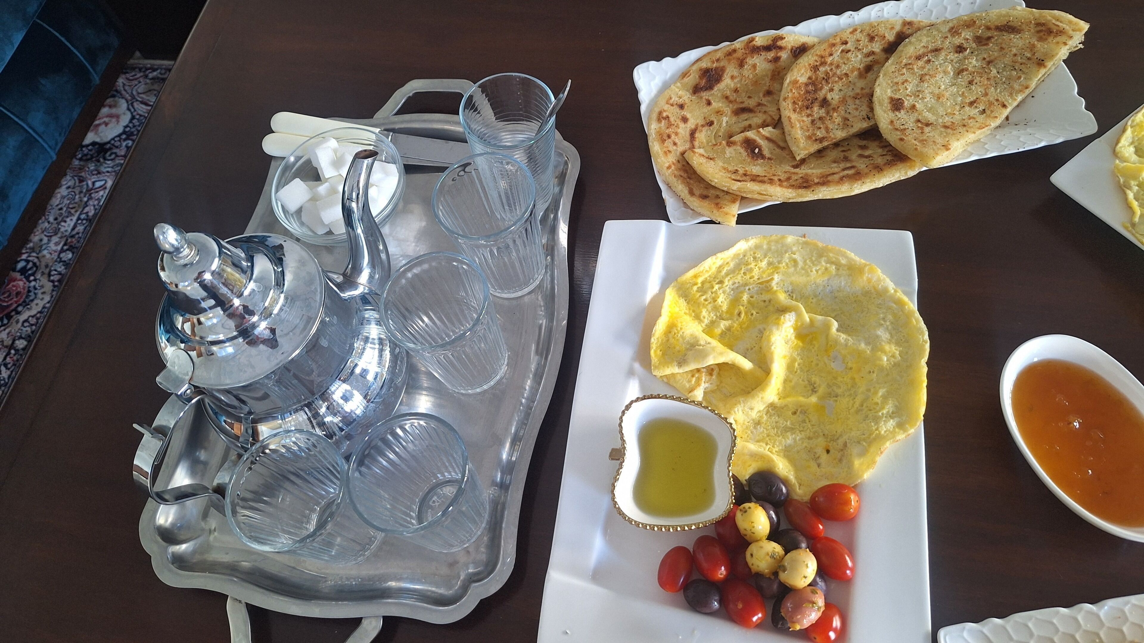A dining table with a teapot, glasses, a plate with eggs, toast, olives, and a small bowl of sauce.