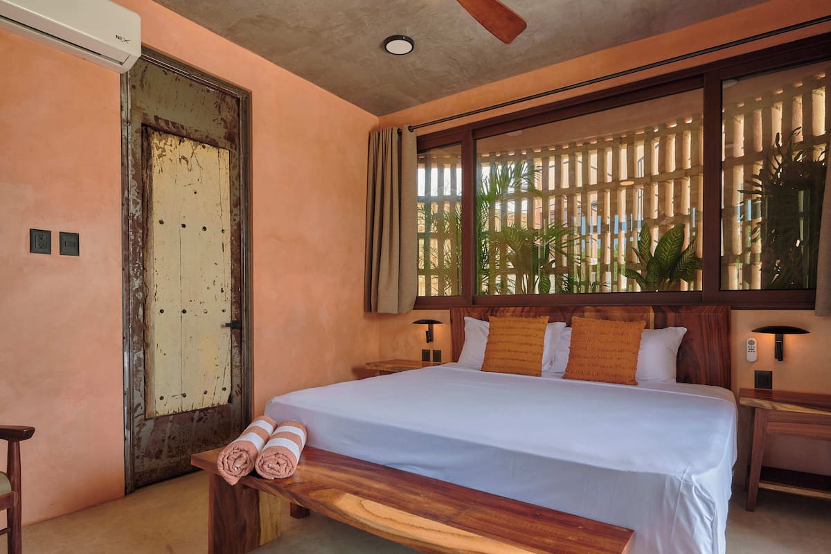 A hotel room with a bed, a wooden bench with towels, a nightstand, a wall-mounted lamp, and a window with a view of greenery.
