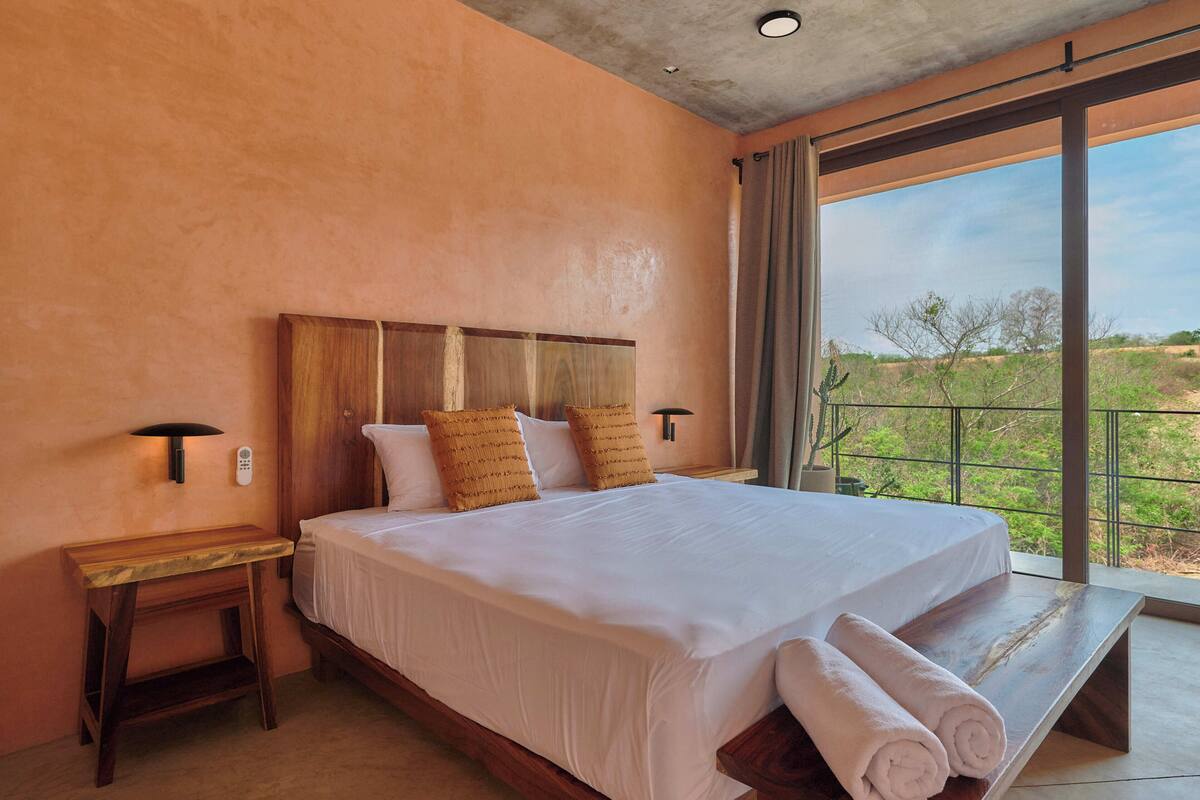 A bedroom with a large bed, wooden headboard, two bedside tables, and a view of a landscape through a large window.