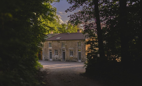 Un bâtiment en pierre avec une allée gravillonnée, entouré d’arbres.
