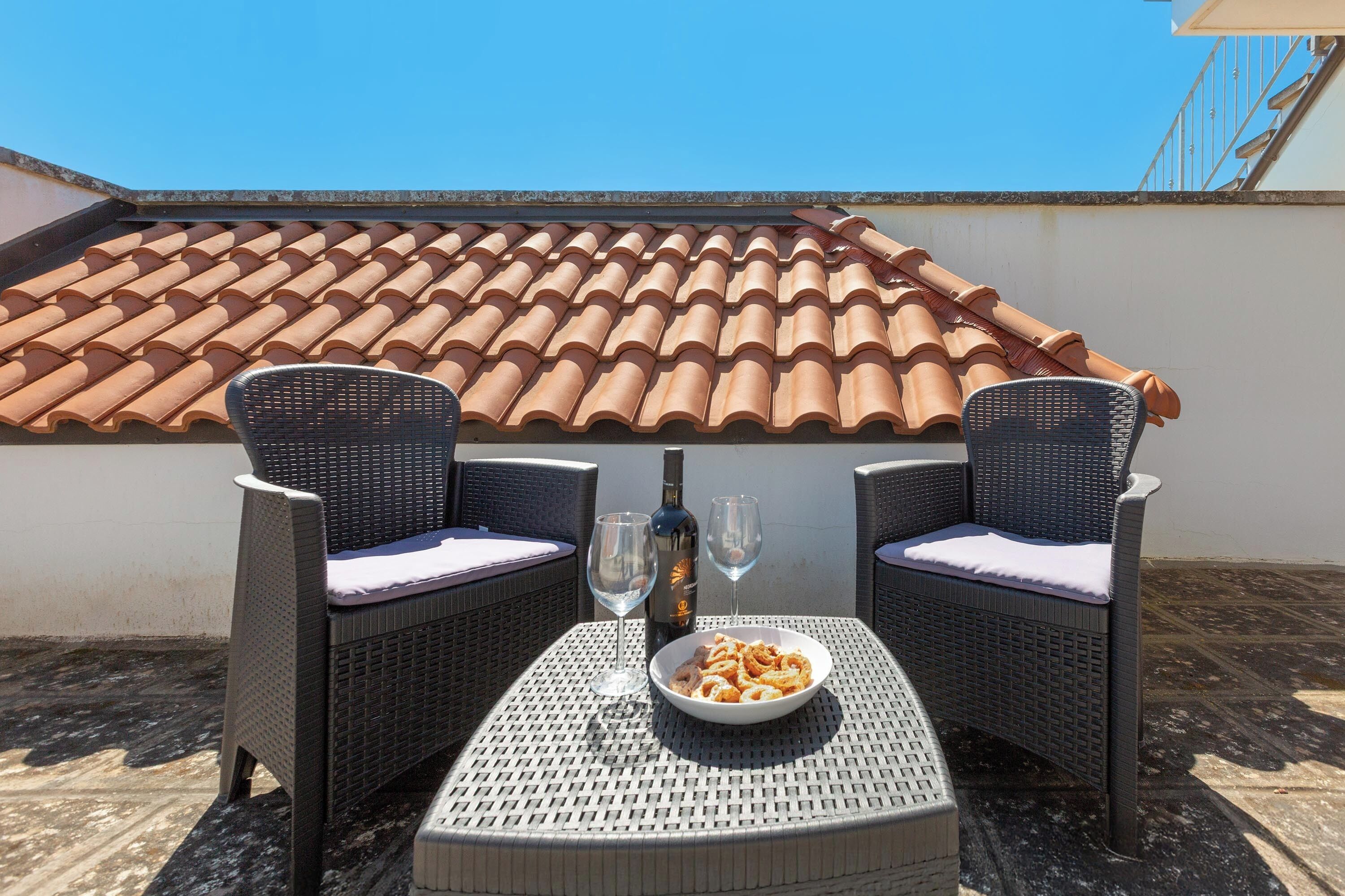 Une terrasse avec deux fauteuils en osier, une table, un bol de collations, une bouteille et deux verres.