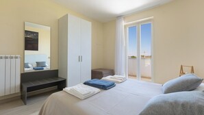 Ein Schlafzimmer mit Bett, einer Sitzbank, einem Kleiderschrank, einem Spiegel und Blick ins Freie.