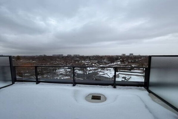 Un balcon enneigé avec une rambarde en métal, une vue sur le paysage urbain et une grille d’évacuation.