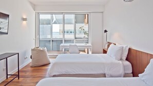 Ett modernt hotellrum med två sängar, ett skrivbord, en stol och ett stort fönster med persienner.
