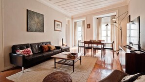 A living room with a black leather sofa, a coffee table, a dining area with a wooden table and chairs, and a TV stand. - Chiado 5 -  Histórico (Lisbon)