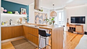 Una cocina moderna con armarios de madera, una isla central, una lámpara colgante y un televisor en la sala de estar contigua.