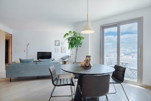 Ein modernes Wohnzimmer mit einer Couch, einem Esstisch mit Stühlen, einem Fernseher und einem großen Fenster mit Blick auf die Stadt.