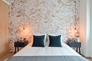 Ein Schlafzimmer mit blumigem Wandpapier, zwei Nachttische mit Lampen, einem Bett mit dunklen Kissen und einem Holzschrank.