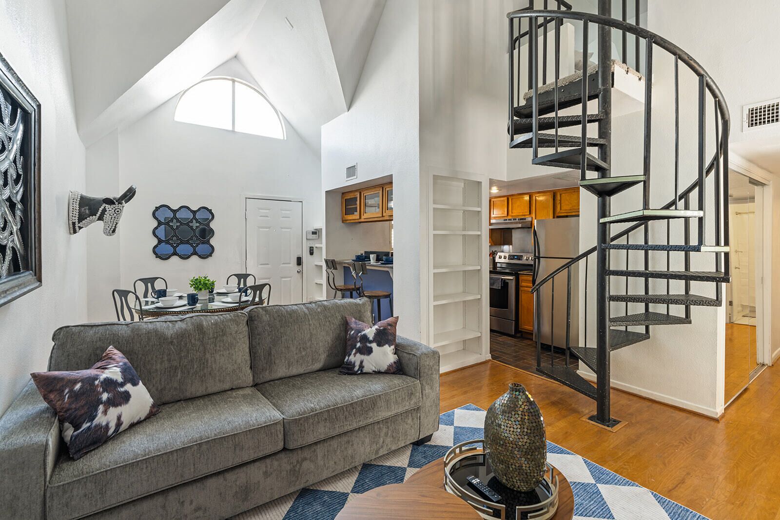 Ein modernes Wohnzimmer mit einem grauen Sofa, einer Wendeltreppe und einer Küche im Hintergrund.