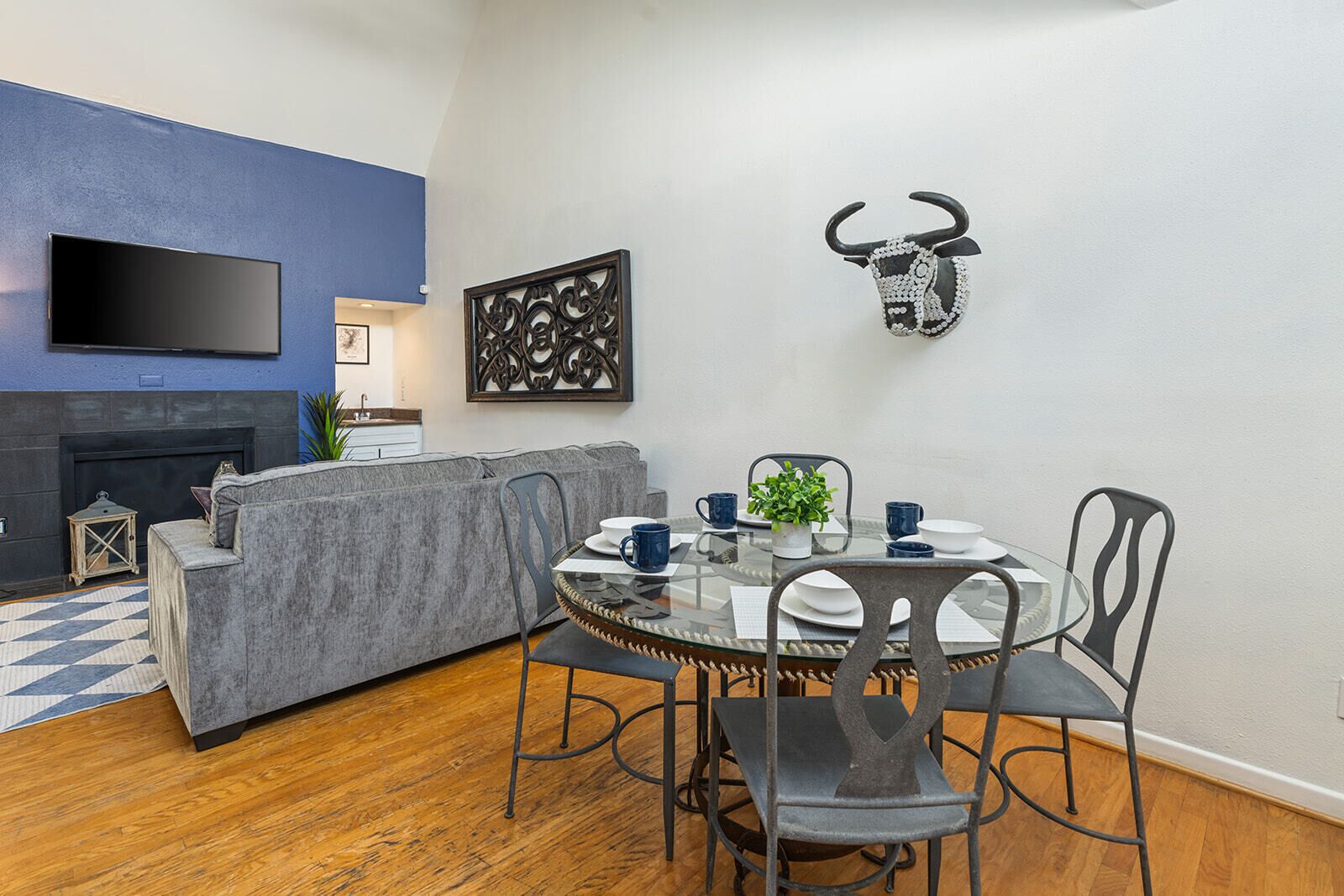 Una sala de estar moderna con un sofá gris, una mesa redonda de vidrio con sillas, un televisor montado, una cabeza de toro decorativa y una chimenea.