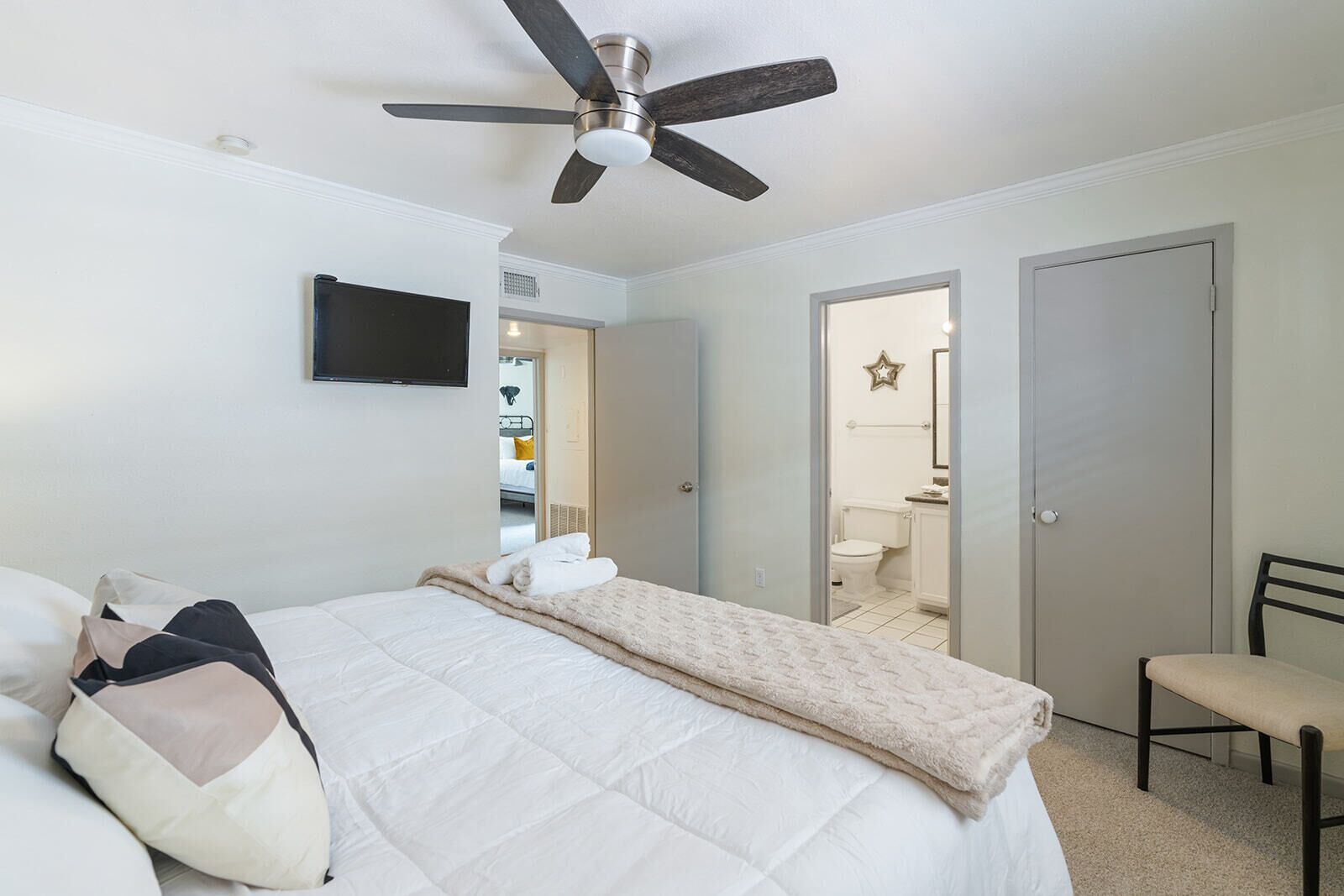 A bedroom with a bed, a ceiling fan, a TV, and a bathroom visible through an open door.