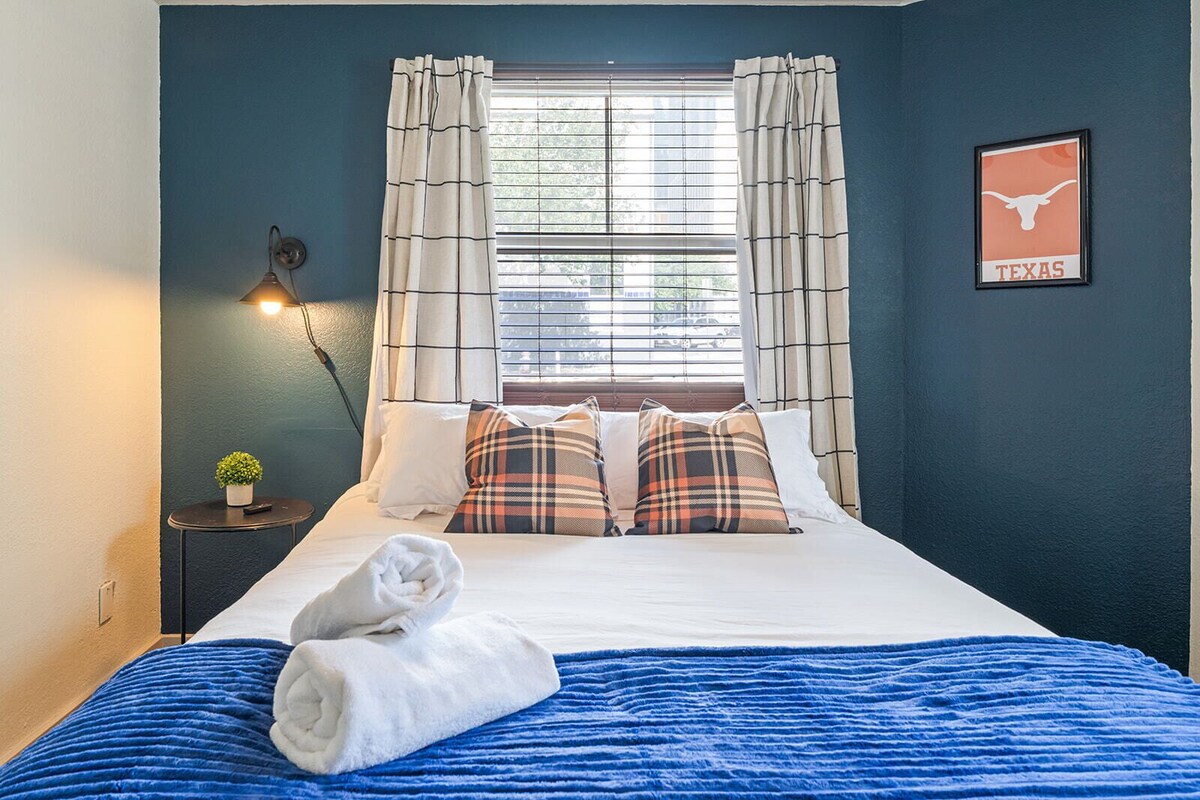 A hotel room with a bed, a window with curtains, a wall-mounted lamp, a small plant, and a Texas-themed wall decoration.