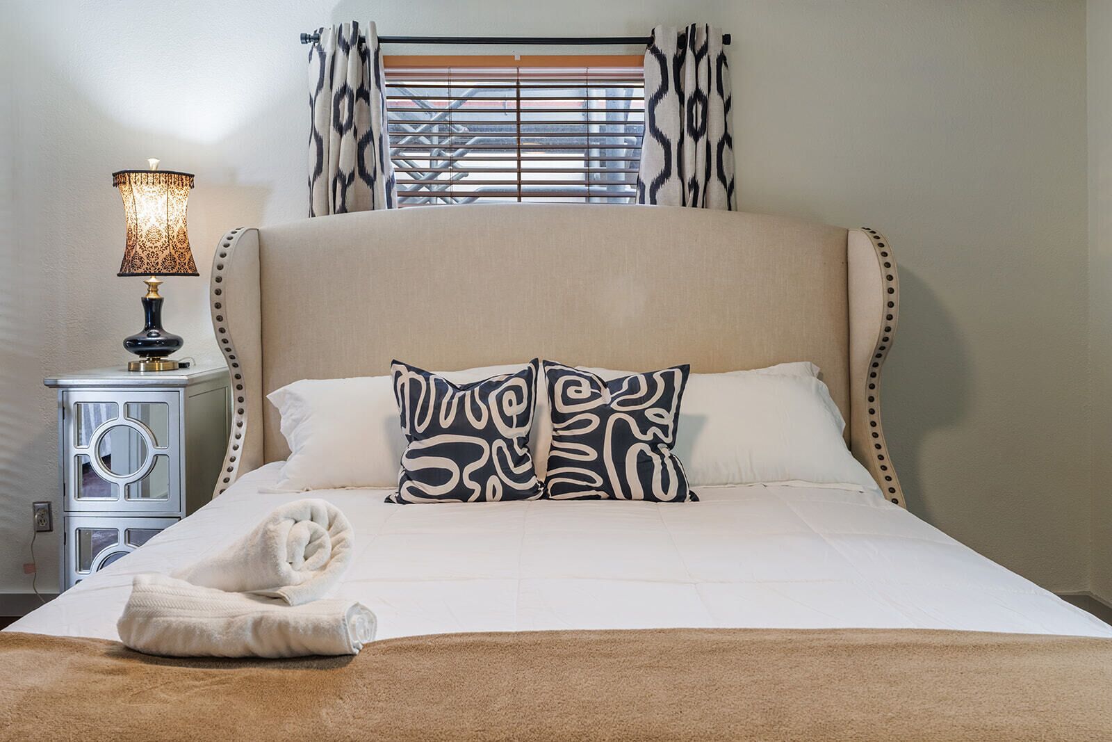 A bed with a headboard, a bedside lamp, a patterned pillow, and a rolled towel on a beige carpet.