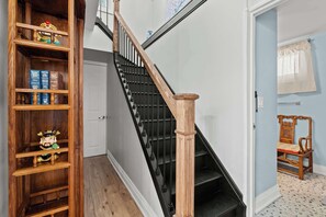 A staircase with a wooden handrail and black balusters, a wooden bookcase with books and a figurine, and a chair in a room with a patterned floor. - The Jade Loft Near Clifton Hill | Soaker Tub 3 Bed (Niagara Falls)