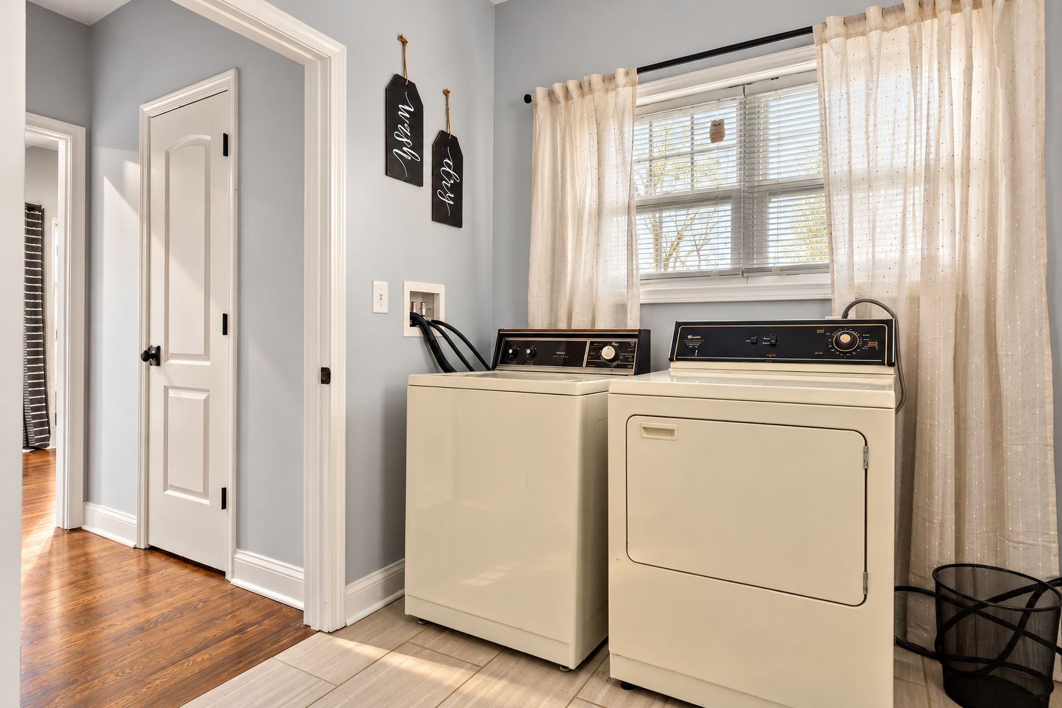 A laundry room with two stacked washers and dryers, a window with sheer curtains, and a door with a white frame.