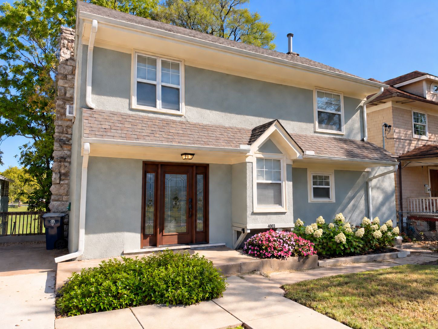 A two-story residential building with a stone facade, a front porch, and a small garden with flowers.