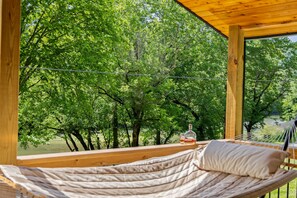 Silla hamaca con un cojín, una botella y vasos sobre una terraza de madera con vista a un lago rodeado de árboles.