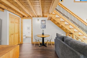 Eine Holztreppe, ein graues Sofa, ein Holzesstisch mit Stühlen und eine weiße Toilette in einem Raum mit Holzbalken an der Decke.