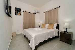 Un dormitorio con cama, cortinas, aire acondicionado, mesita de noche y televisor montado en la pared.