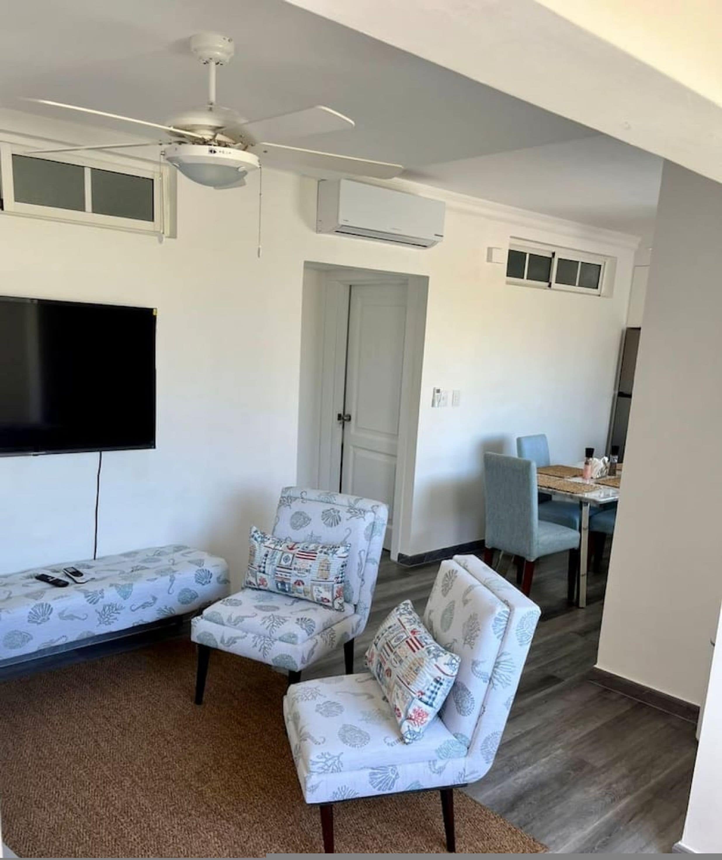 A living room with a sofa, two armchairs, a ceiling fan, a flat-screen TV, and a dining area with a table and chairs.