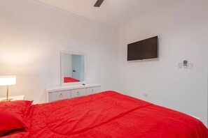 Un dormitorio con cama, mesita de noche con lámpara, cómoda, espejo y televisor montado en la pared.