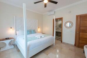 Un dormitorio con cama con dosel, una mesita de noche, ventilador de techo, espejo y una puerta que da a un baño.