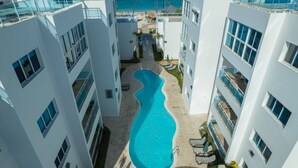 A modern hotel with a central swimming pool surrounded by white buildings, balconies, and a view of the beach. - Beachfront Condo 2 Pools Private Beach 8 Guest (Punta Cana)