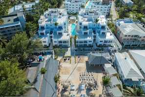 Vue aérienne d’un complexe hôtelier comprenant plusieurs bâtiments, une piscine centrale et une zone de plage avec des transats.