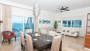 A modern living room with a dining area, large windows, a ceiling fan, and a view of the ocean. - Beachfront Condo 2 Pools Private Beach 8 Guest (Punta Cana)