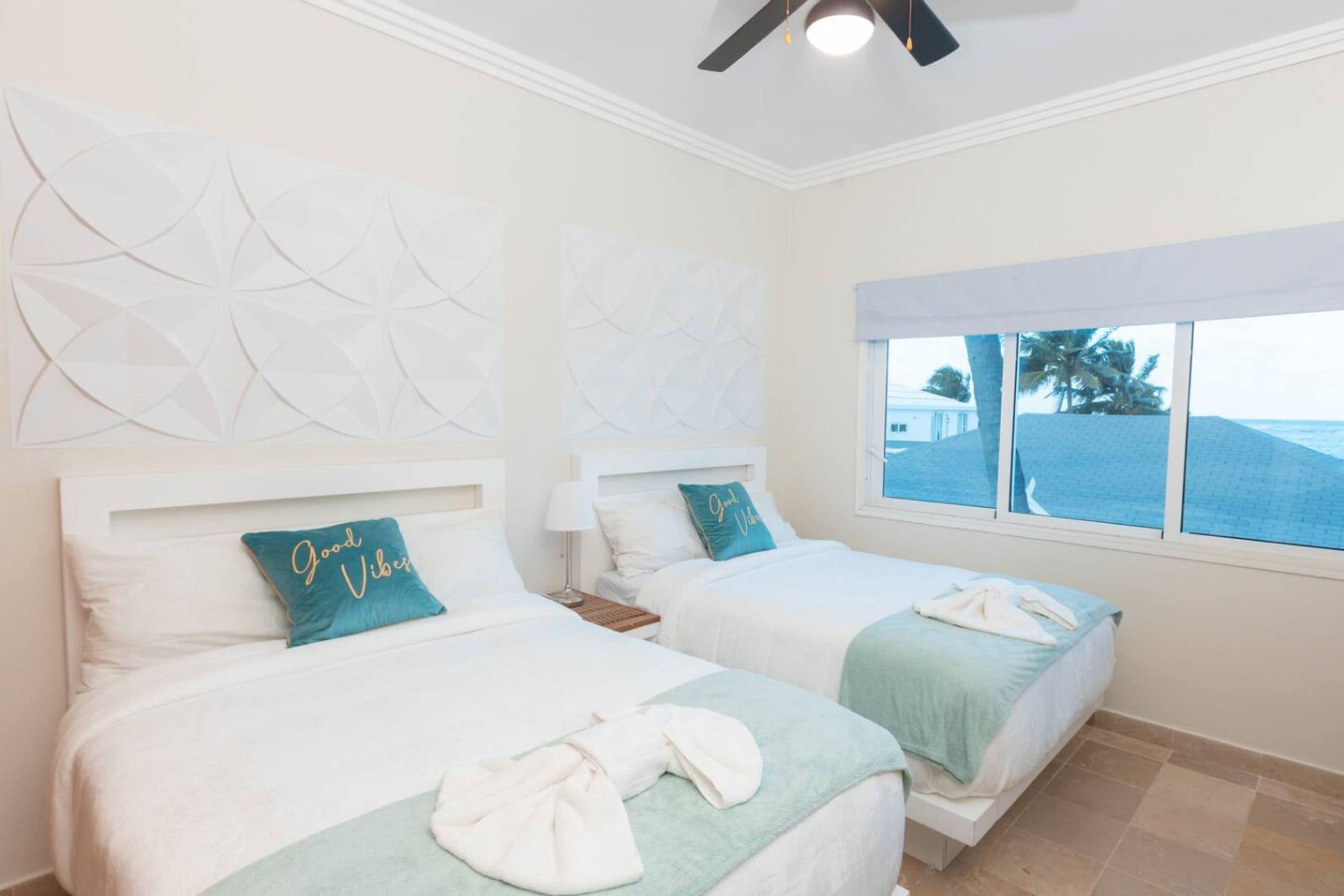 A hotel room with two beds, a ceiling fan, a window with a view of the ocean, and decorative wall panels.