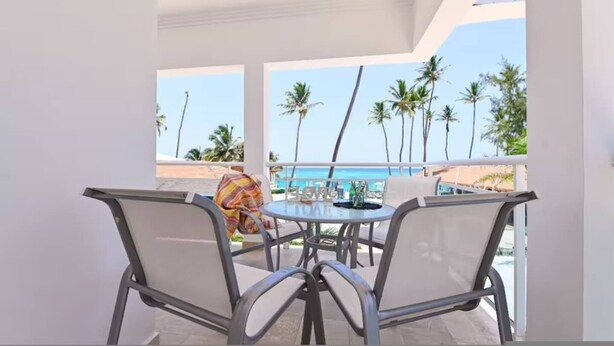 A balcony with a table and chairs, overlooking a beach with palm trees and the ocean. - Beachfront Condo 2 Pools Private Beach 8 Guest (Punta Cana)