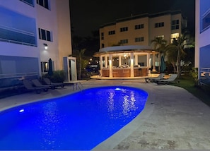 Une piscine avec des chaises longues, un espace bar et un bâtiment à plusieurs étages.