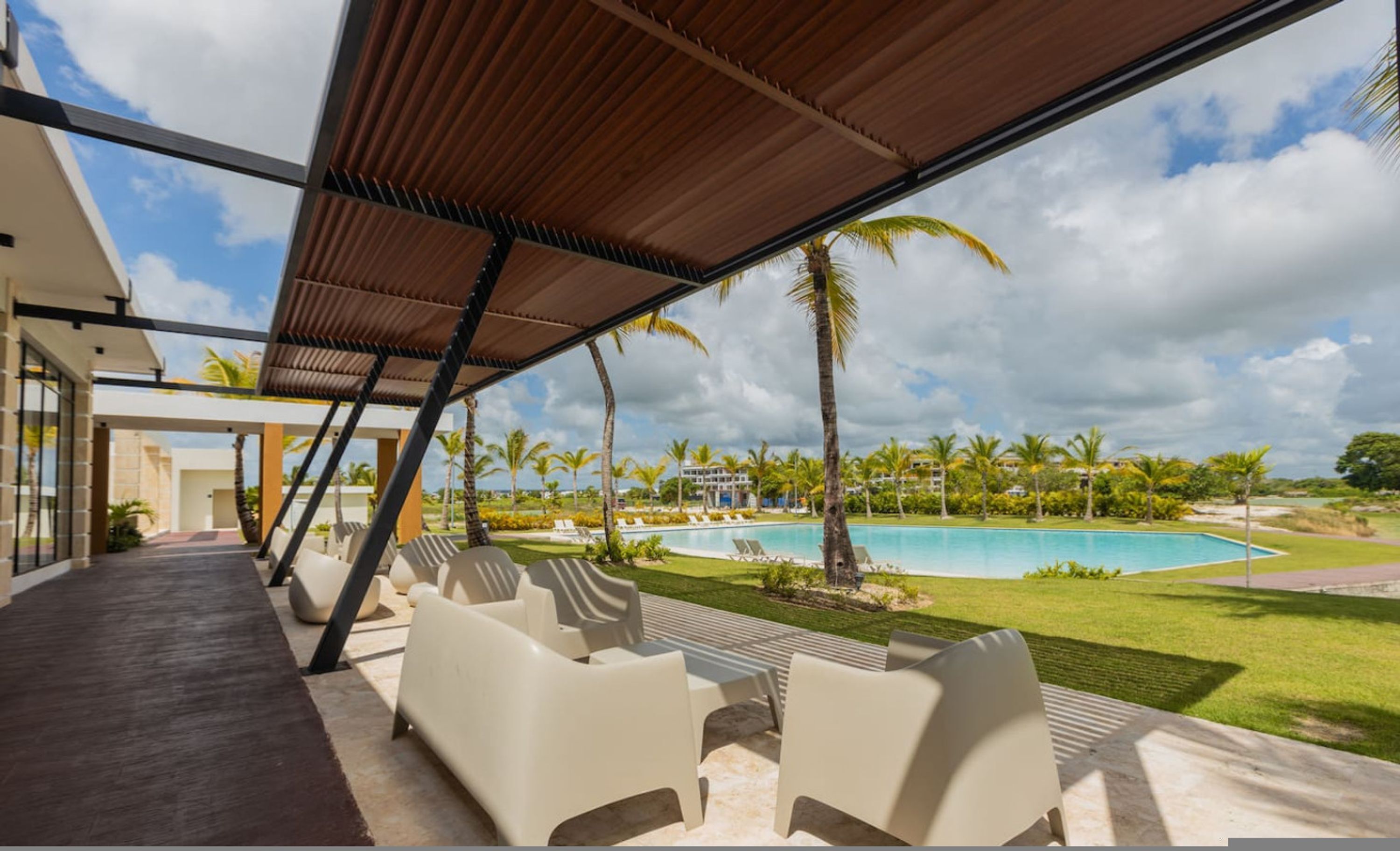 A poolside area with lounge chairs, a covered patio, and a view of a swimming pool and palm trees.