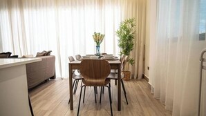 Una sala de estar moderna con un sofá, una mesa de comedor, sillas, una planta en maceta y un amplio ventanal con cortinas traslúcidas.